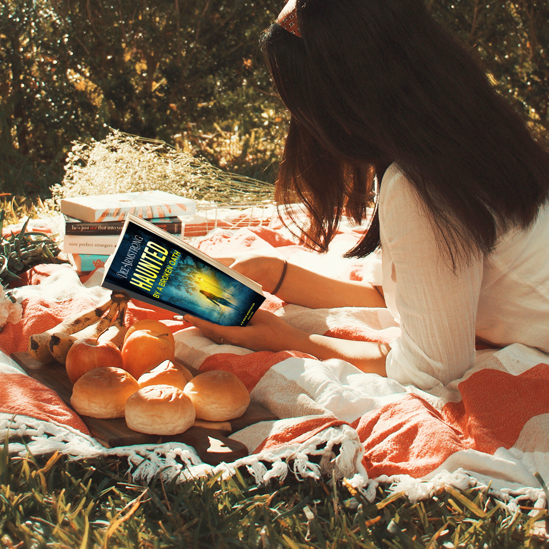 Person reading a Haunted by a Broken Oath by Dee Armstrong outdoors with a blanket and food items