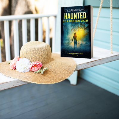 Book titled 'Haunted by a Broken Oath' by Dee Armstrong on a swing with a sun hat and flowers.