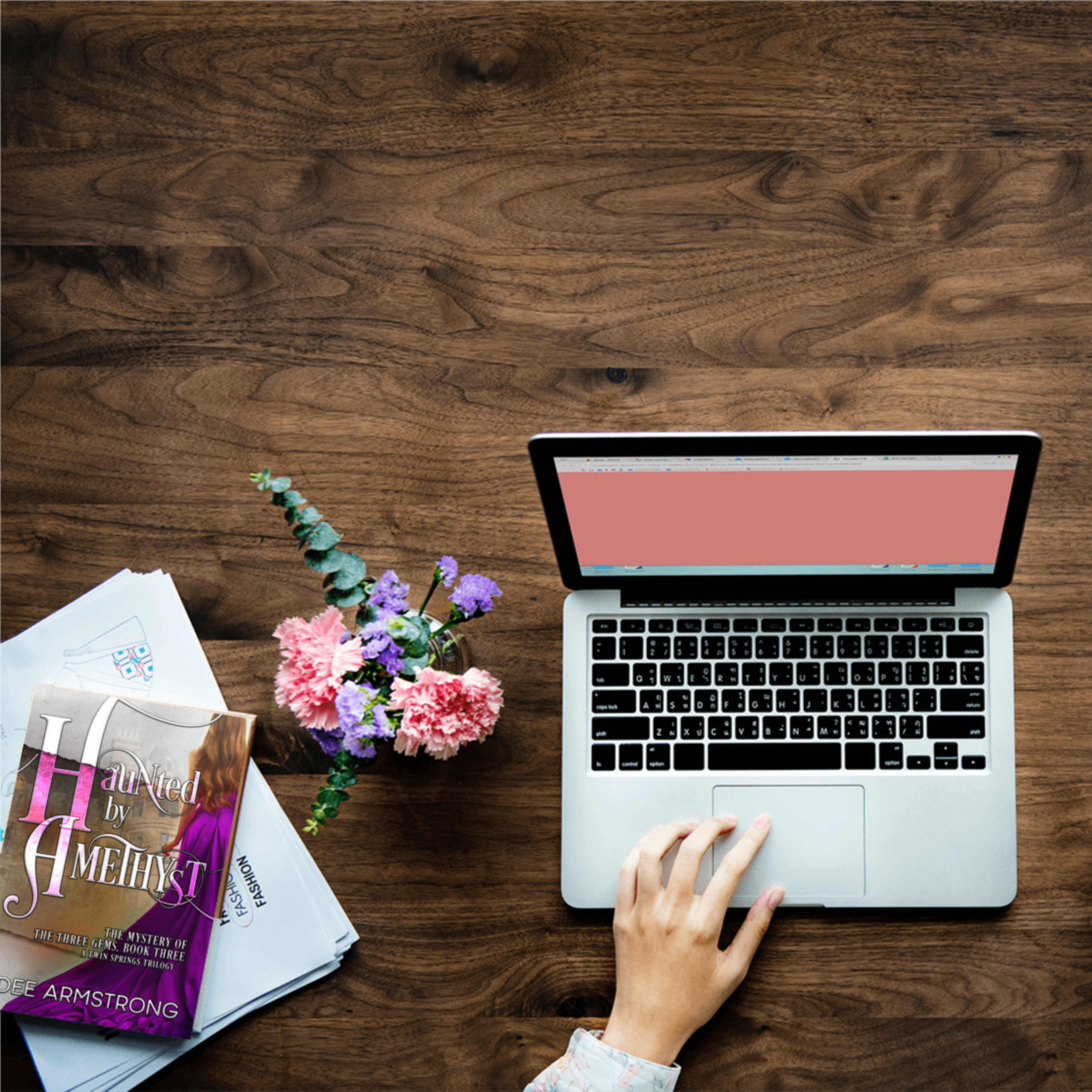 Laptop on a wooden desk with the book Haunted by Amethyst and flowers