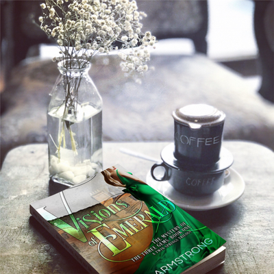 Book titled 'Visions of Emerald' by Dee Armstrong with a cup of coffee and a vase on a table.